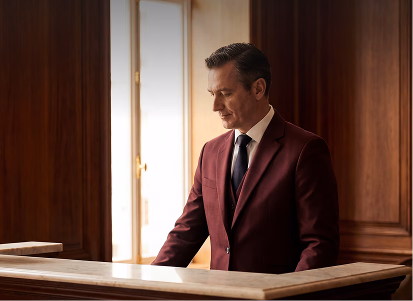 Man in a burgundy suit, white shirt, and black tie standing behind a polished wooden counter with marble top, looking down to the right in a softly lit room with wooden panel walls and a window in the background.
