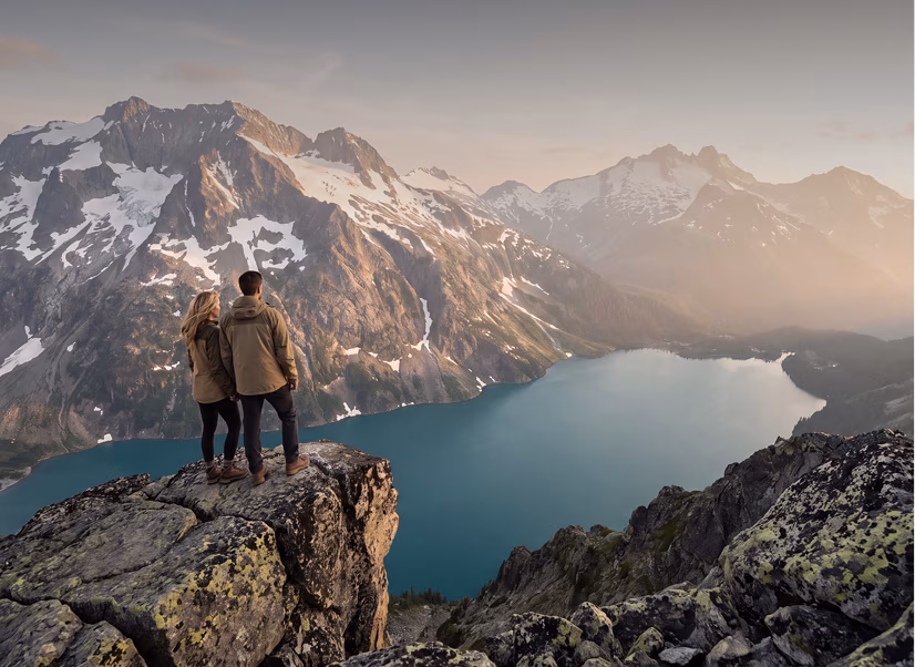 A man and woman wearing jackets stand on a rocky cliff overlooking a blue mountain lake surrounded by snow-capped peaks under a cloudy sky at sunset.