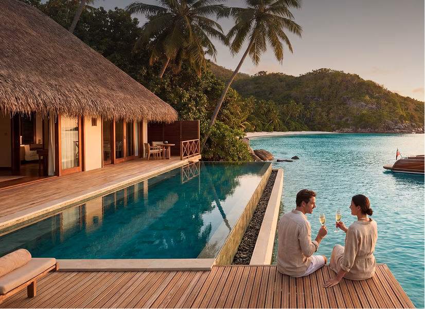 Couple sitting on a wooden deck by an infinity pool next to a tropical thatched-roof bungalow, clinking glasses overlooking clear turquoise water and lush green hills at sunset.