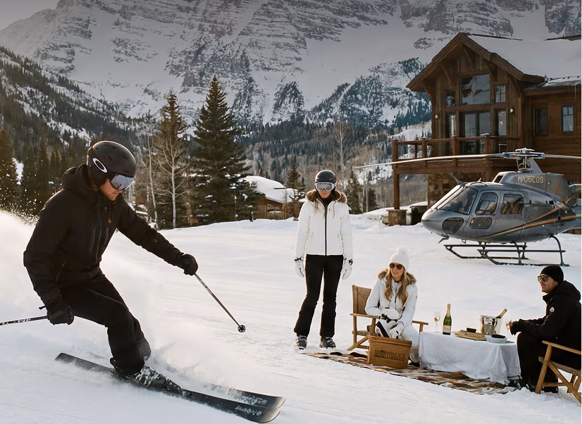 A skier in black gear and helmet skiing down a snowy slope near three people by a picnic setup with drinks and food; a wooden cabin and a helicopter are in the background with tall pine trees and snow-covered mountains behind.