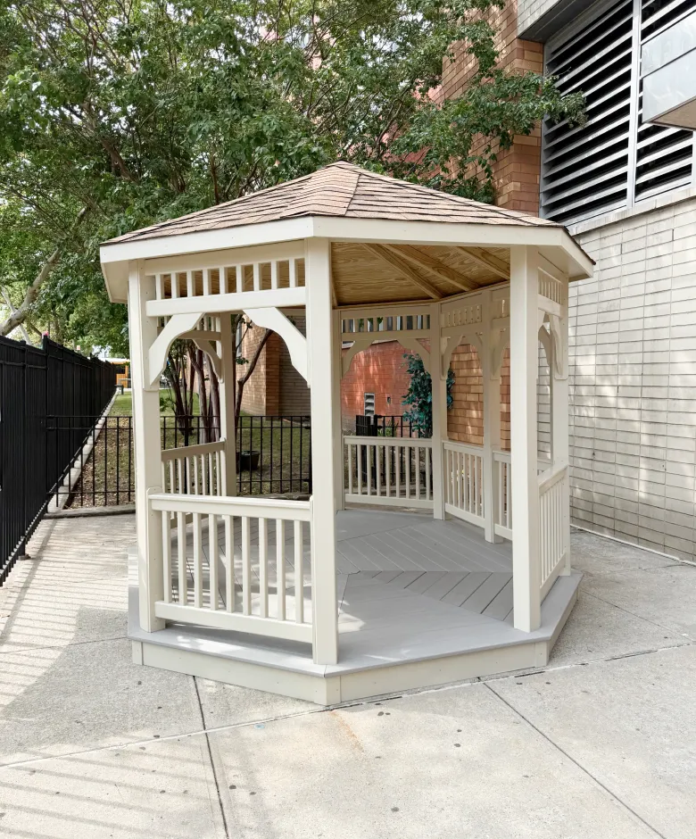 A light-colored, octagonal wooden gazebo sitting on a concrete patio next to a brick building.