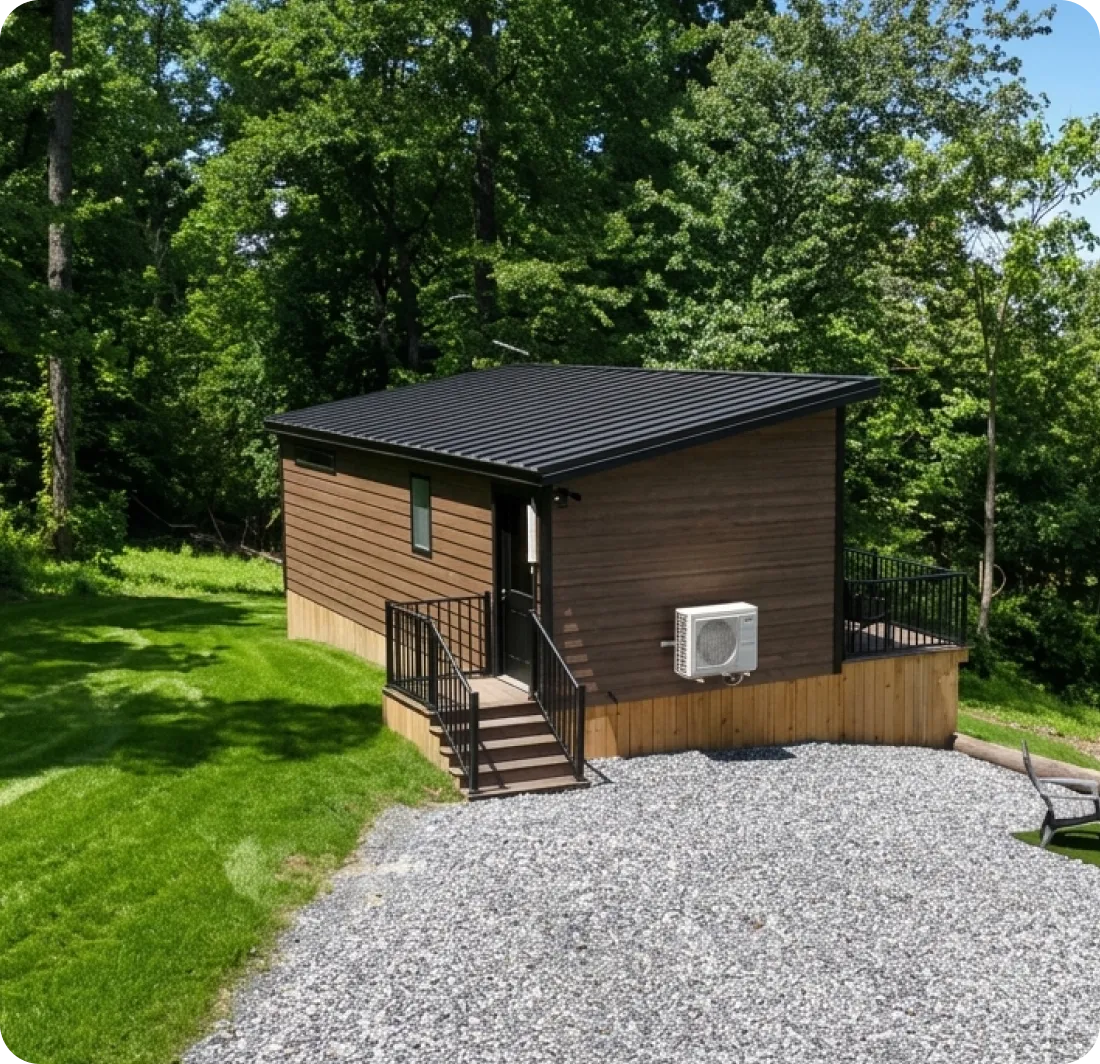 A small, modern cabin with brown horizontal siding, a black shed roof, and a small front deck, situated on a gravel lot next to a grassy lawn.