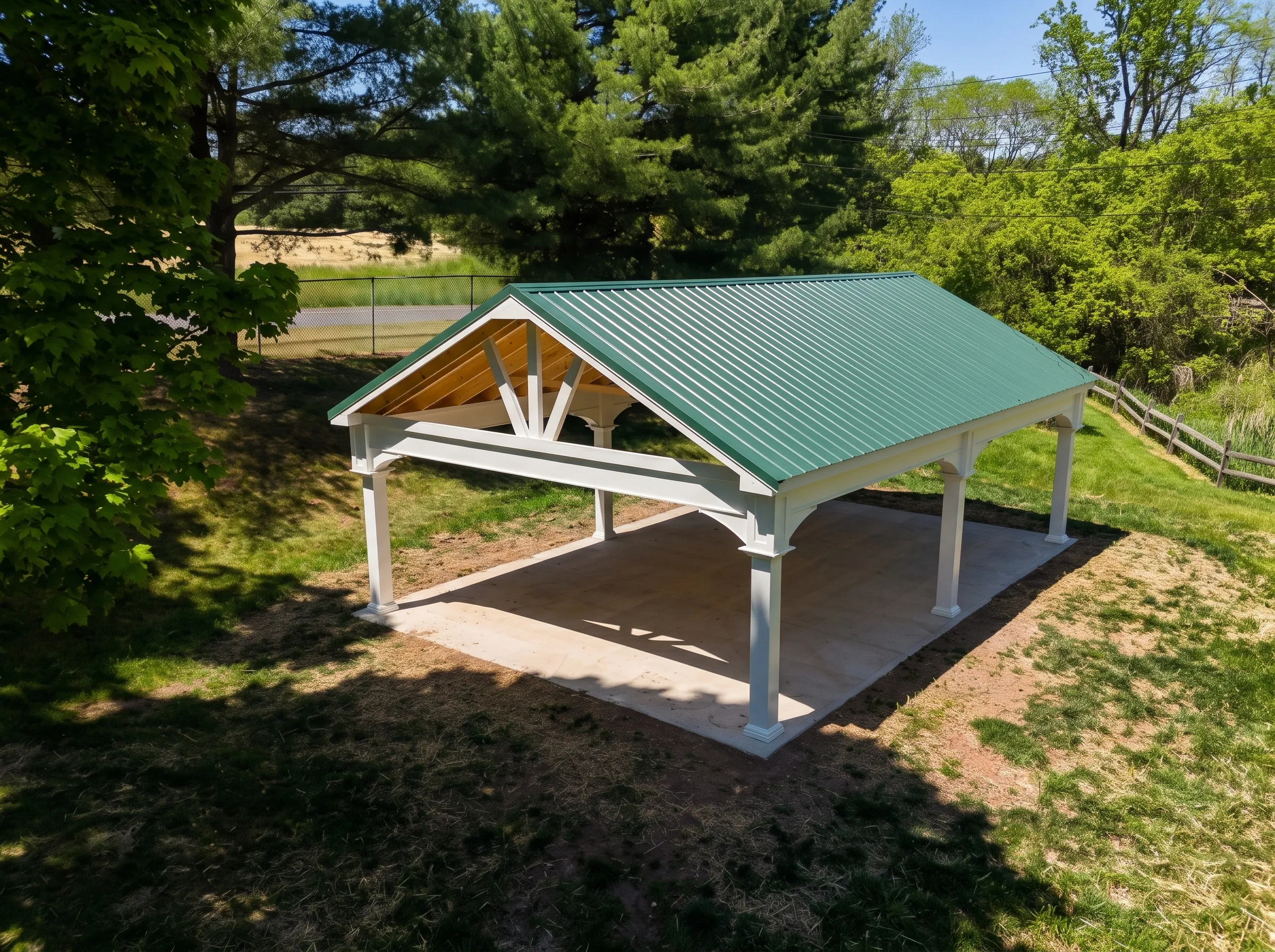 A white open-air pavilion with a green metal gable roof situated on a concrete slab in a grassy yard.