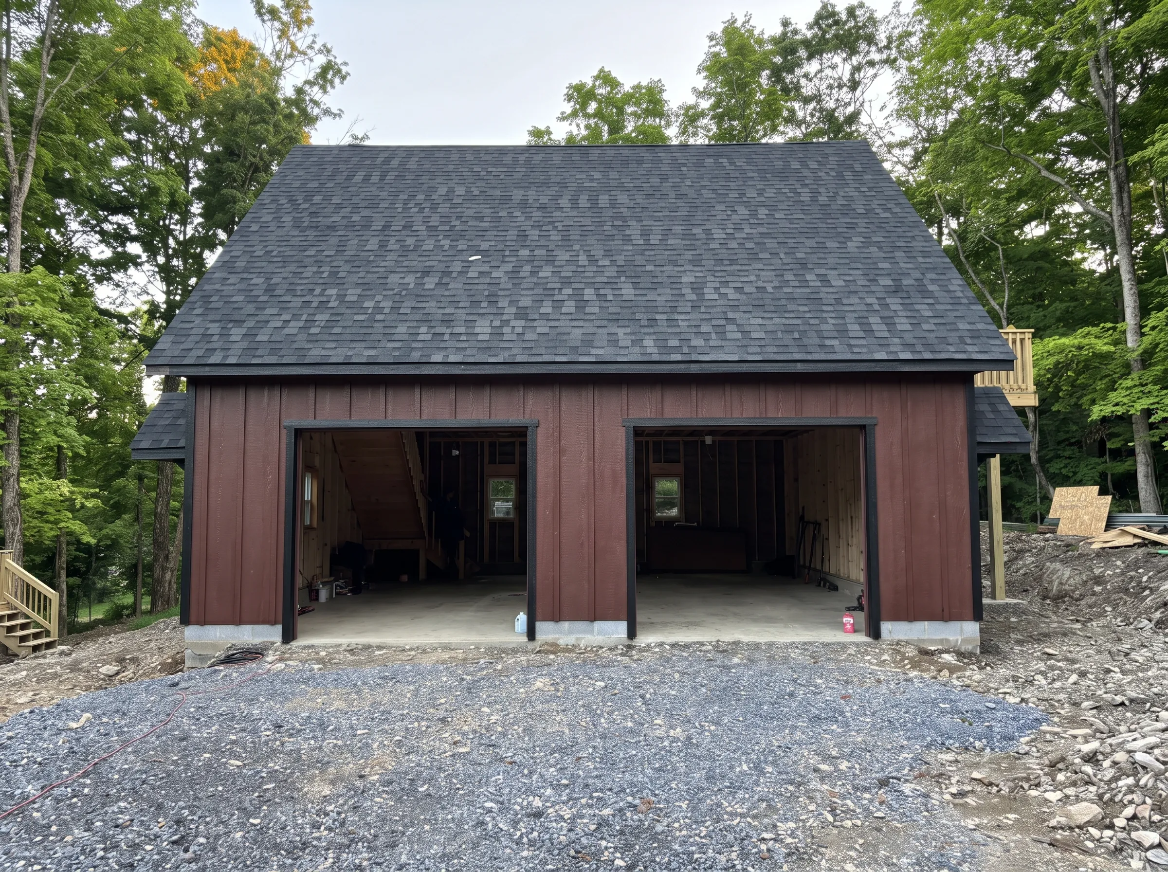A two-bay garage under construction with brown vertical siding, a dark shingled roof, and an unfinished gravel driveway.