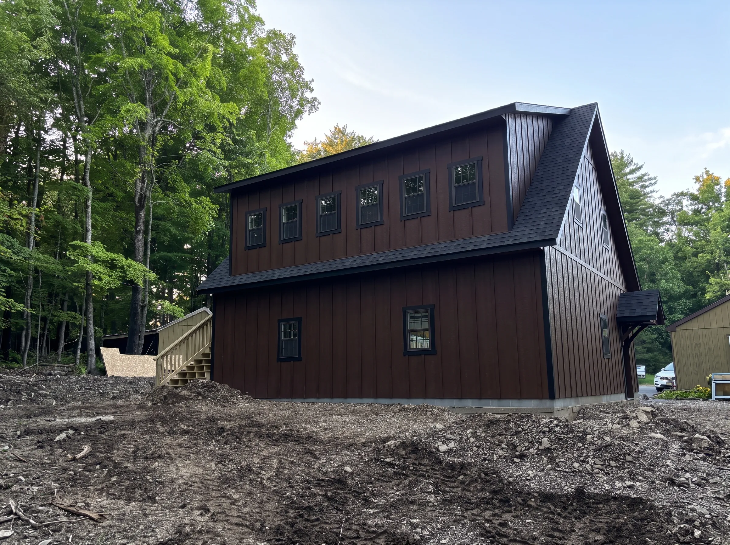 A two-story brown metal barn-style building with black trim, situated on a dirt lot next to a forest.