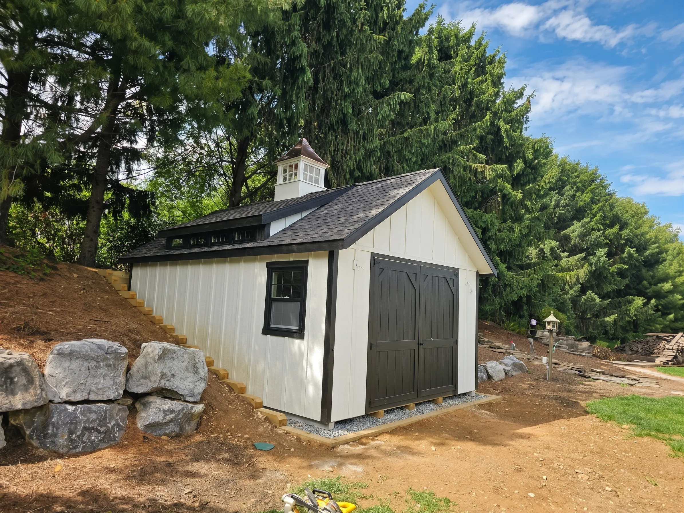 A white storage shed with black trim and double doors, situated on a sloped lot next to large gray boulders and evergreen trees.