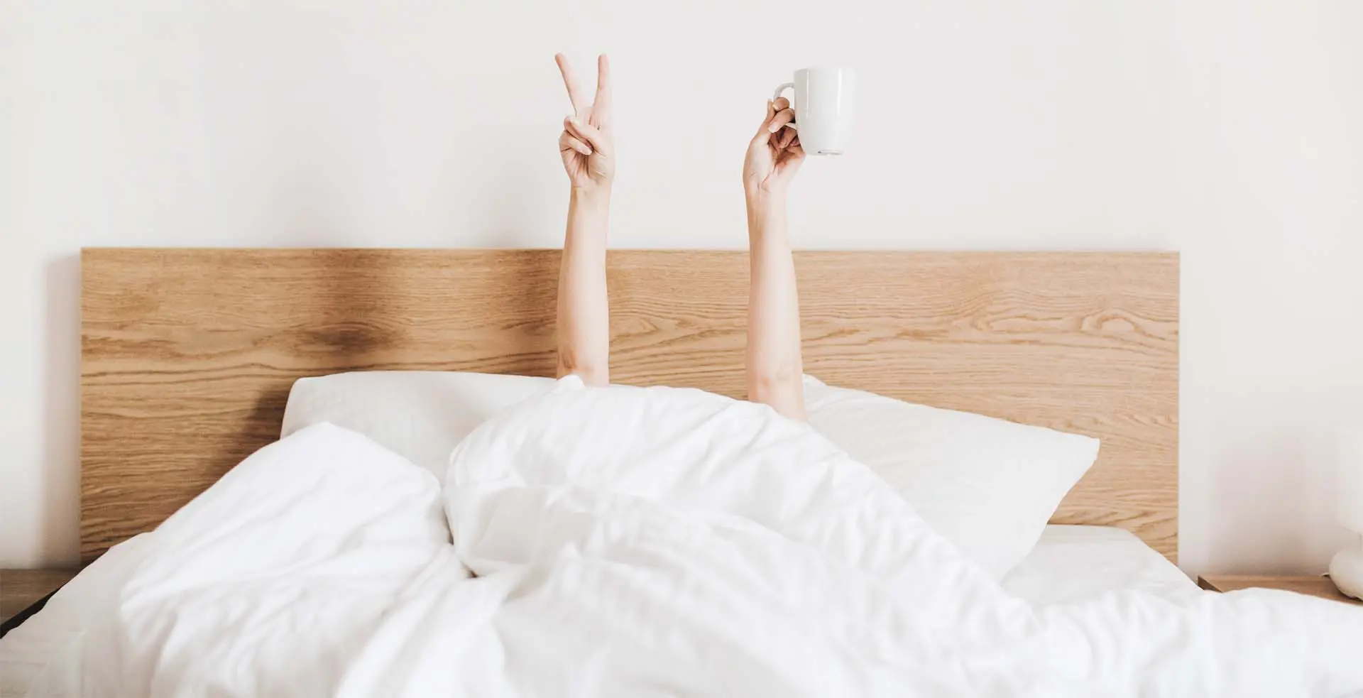 Person liegt unter weißer Bettdecke auf einem Bett mit Holz-Kopfteil und hält eine Hand mit Peace-Zeichen und eine Hand mit einer weißen Tasse hoch.