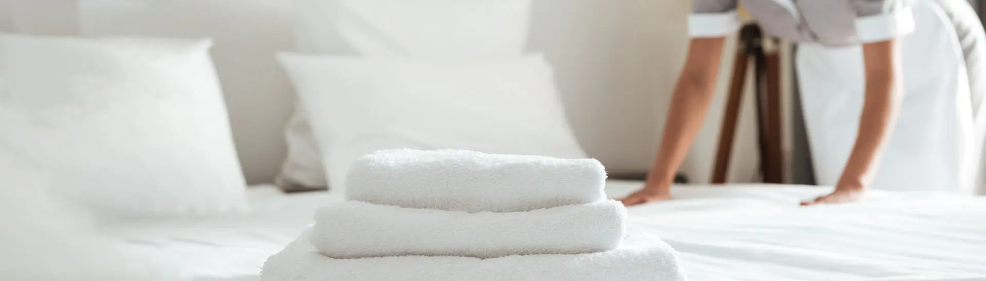 Gestapelte, saubere weiße Handtücher liegen auf einem frisch gemachten Bett mit weißen Kissen, während eine Person im weißen Dienstkleid im Hintergrund die Bettdecke richtet.