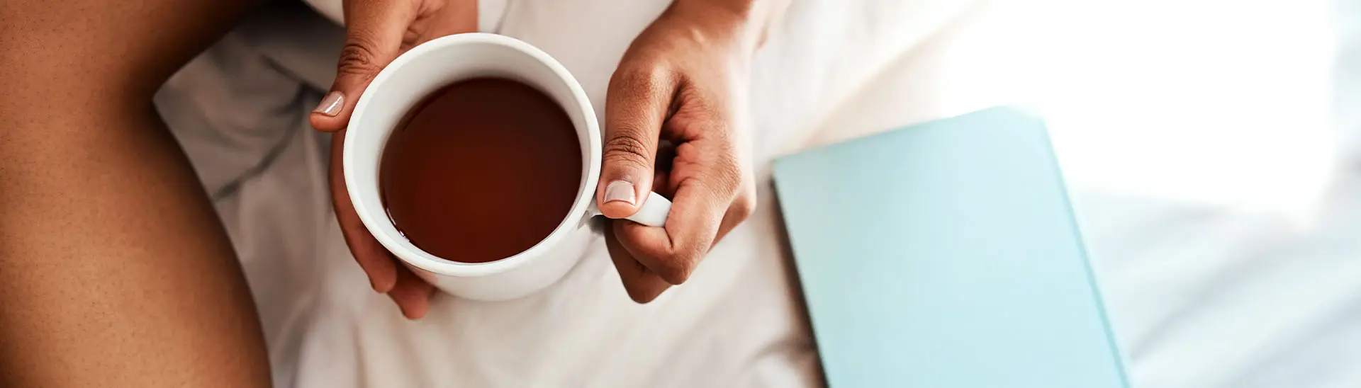 Eine Person hält eine weiße Tasse mit Tee oder Kaffee, sitzend auf einem Bett mit weißen Laken und einem blauen Buch oder Notizbuch daneben.