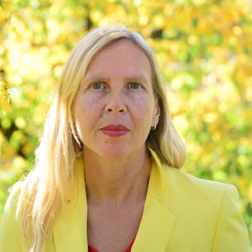 Porträt einer Frau mit langen blonden Haaren und neutralem Gesichtsausdruck, die eine gelbe Jacke trägt, vor einem verschwommenen, grünen Hintergrund.