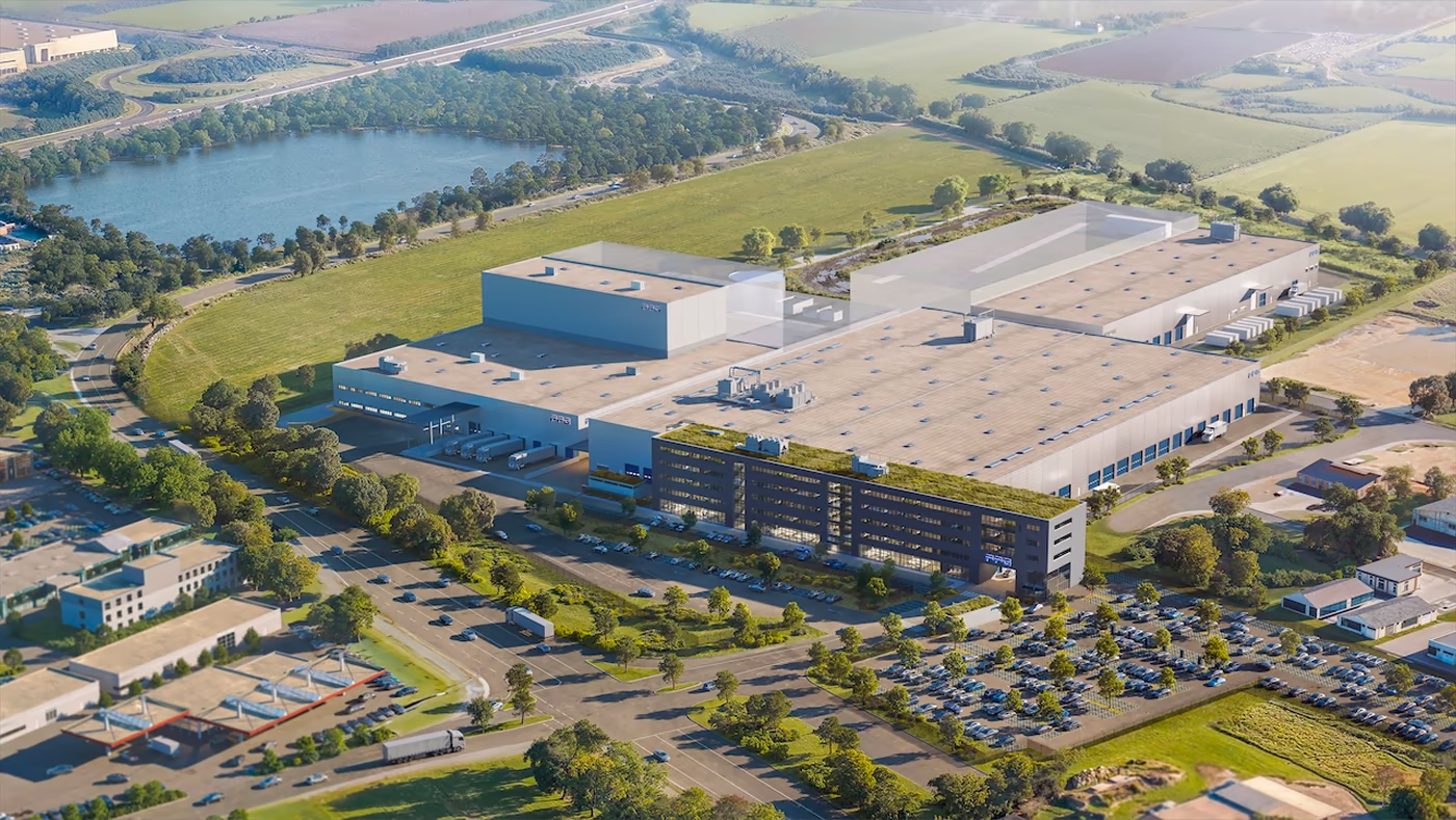 Luftaufnahme eines großen Industriekomplexes mit mehreren verbundenen Gebäuden und Parkplätzen, umgeben von grünen Feldern, Bäumen und einem nahegelegenen See.