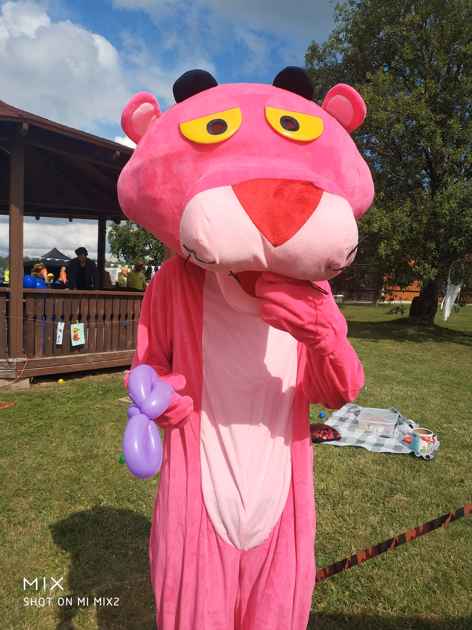 Person dressed in a large pink panther costume standing on grass outdoors near a wooden pavilion, holding a purple balloon animal in one hand, with the other hand near the mouth. People and trees are visible in the background under a partly cloudy sky.