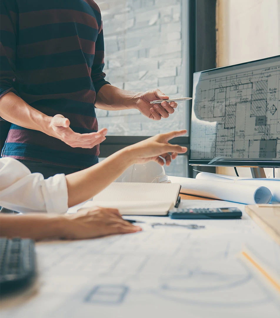 Zwei Personen besprechen Architekturpläne, einer zeigt mit einem Stift auf einen digitalen Bauplan auf einem Monitor, während der andere auf einen Papierplan zeigt.