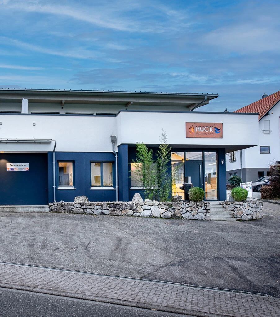 Modernes Firmengebäude mit weißer und dunkelblauer Fassade, Steinmauer und kleiner Treppe vorne, einem Schild mit dem Namen 'HUCK' über dem Eingang und einem Parkplatz im Vordergrund unter bewölktem Himmel.
