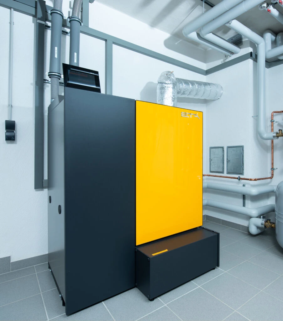 Moderne Heizungsanlage mit schwarzem und gelbem Gehäuse in einem Technikraum mit Rohrleitungen an den Wänden.