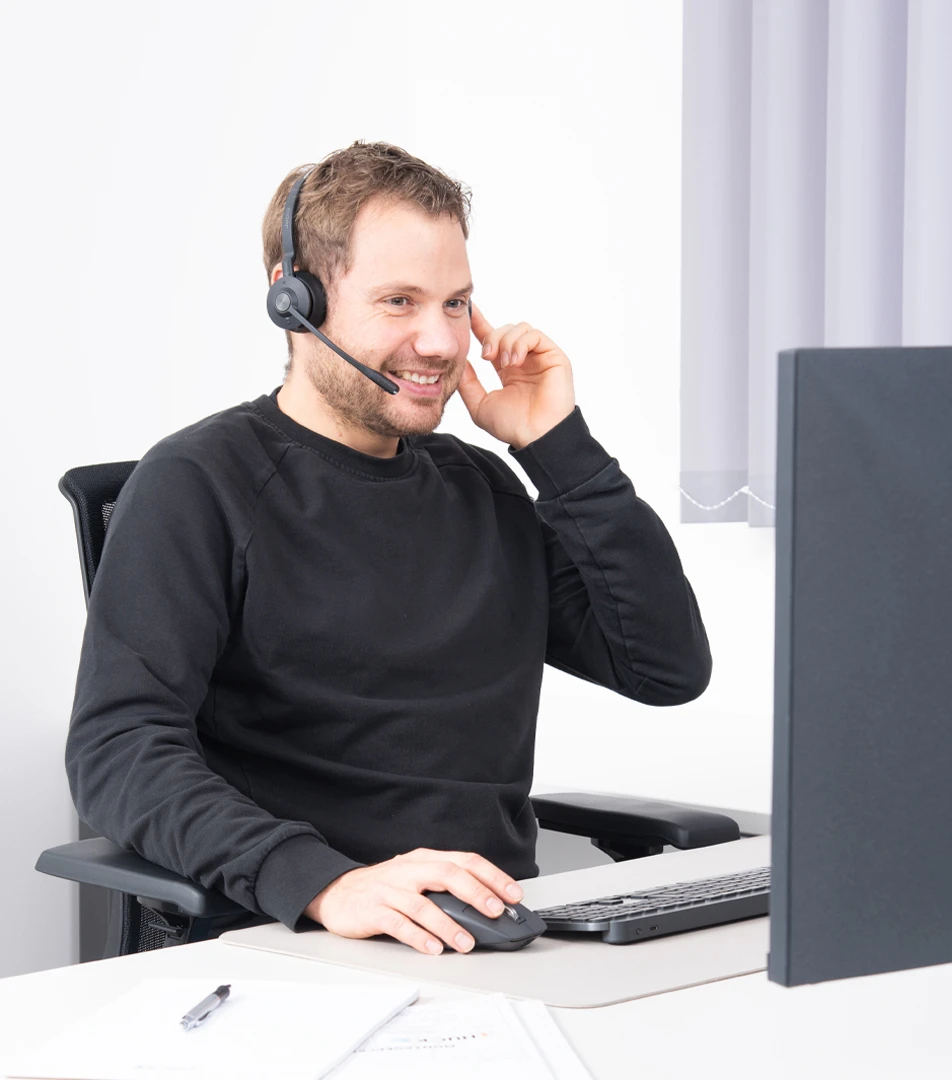 Junger Mann in schwarzem Pullover sitzt an einem Schreibtisch vor einem Computerbildschirm und trägt ein Headset, während er eine Computermaus benutzt und lächelt.