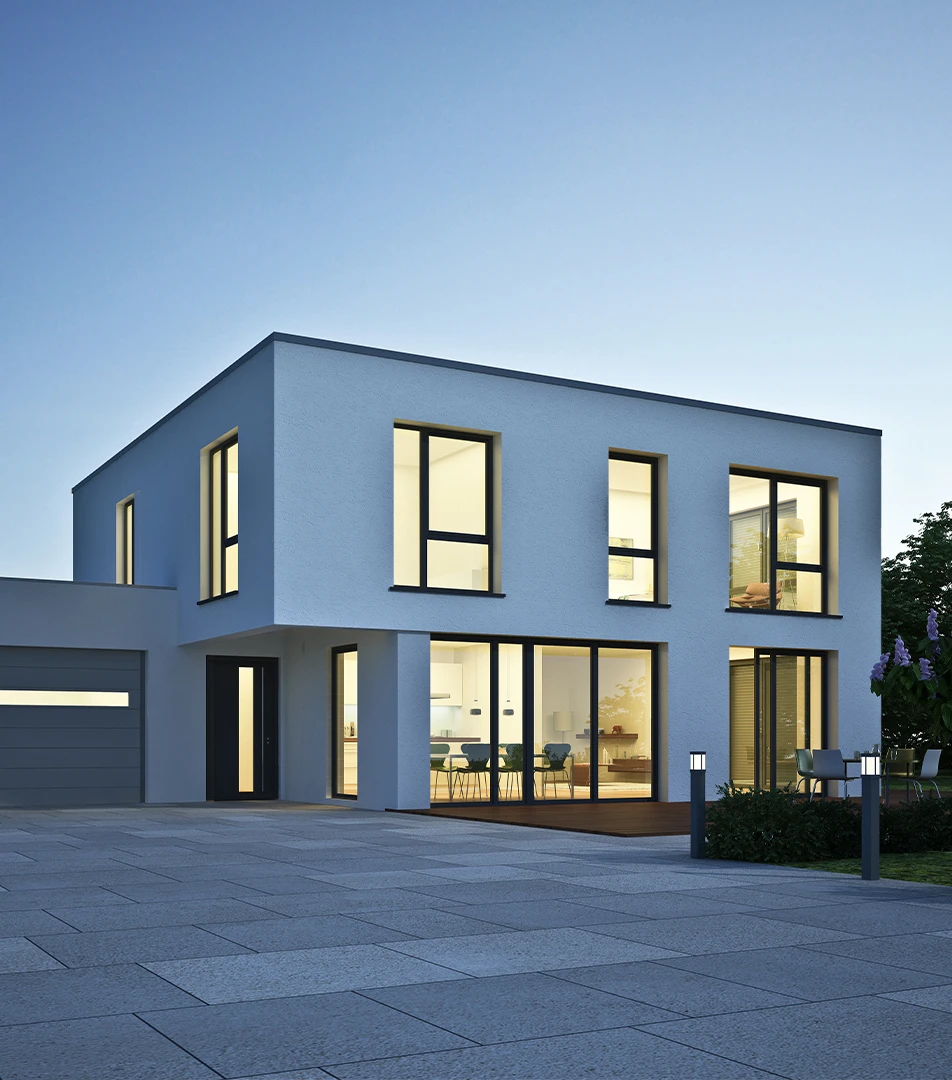 Modern zweistöckiges Haus mit weißen Wänden und großen Fenstern, beleuchtet von innen in der Abenddämmerung, mit einer gepflasterten Einfahrt und Gartenbeleuchtung.