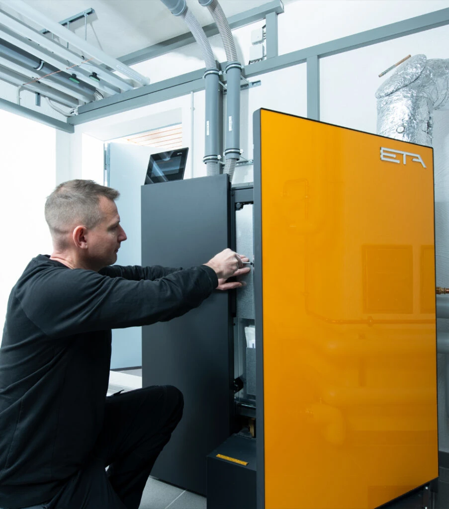 Ein Mann kniet neben einem großen orangefarbenen Heizkessel oder Industriegerät und justiert oder repariert dessen schwarze Seitenklappe in einem technischen Raum mit Rohren an der Decke.