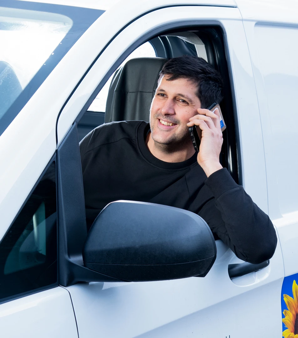 Lächelnder Mann mit schwarzem Pullover, der aus dem Fenster eines weißen Lieferwagens schaut und mit dem Handy telefoniert.