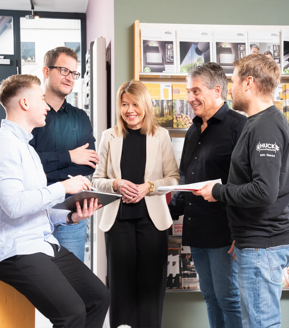 Fünf Personen in einem hellen Büro stehen in lockerer Gesprächsrunde, eine Person sitzt mit einem Notizblock auf einem Hocker; im Hintergrund Bücher- und Prospektständer.