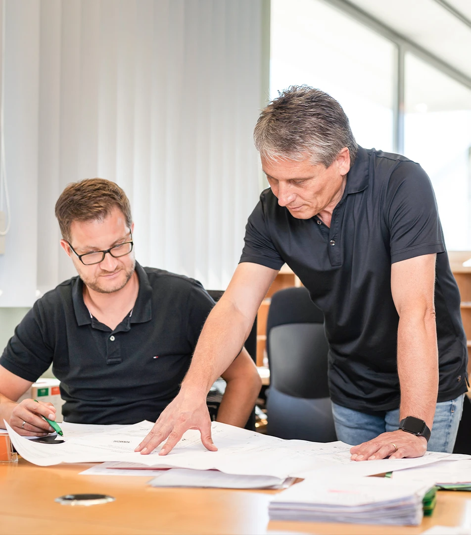 Zwei Männer in lässiger Kleidung besprechen und prüfen Pläne auf einem Schreibtisch in einem hellen Büro.