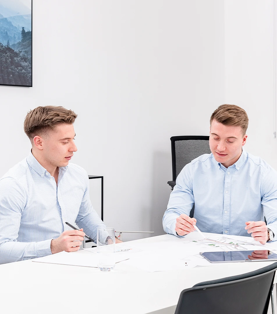 Zwei junge Männer in hellblauen Hemden sitzen an einem weißen Tisch in einem modernen Büro und arbeiten an Dokumenten mit Stiften, vor ihnen liegt ein Tablet.