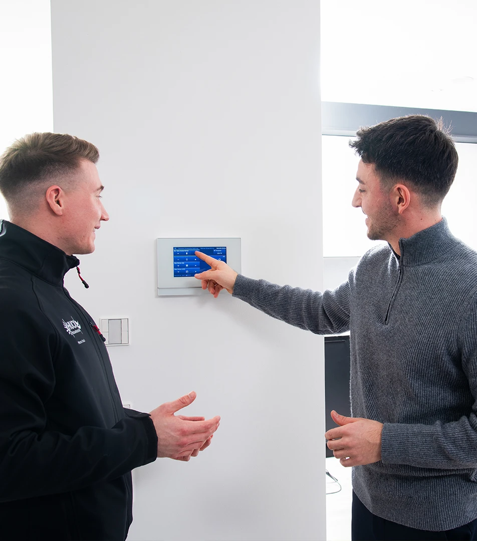 Zwei Männer stehen vor einer Wand, einer zeigt auf ein Wandthermostat mit blauem Display, der andere hört zu und gestikuliert mit den Händen.