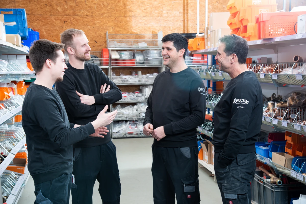 Vier Männer in schwarzer Arbeitskleidung führen in einem Lagergespräch, umgeben von Regalen mit verschiedenen Installationsmaterialien wie Rohren und Behältern.