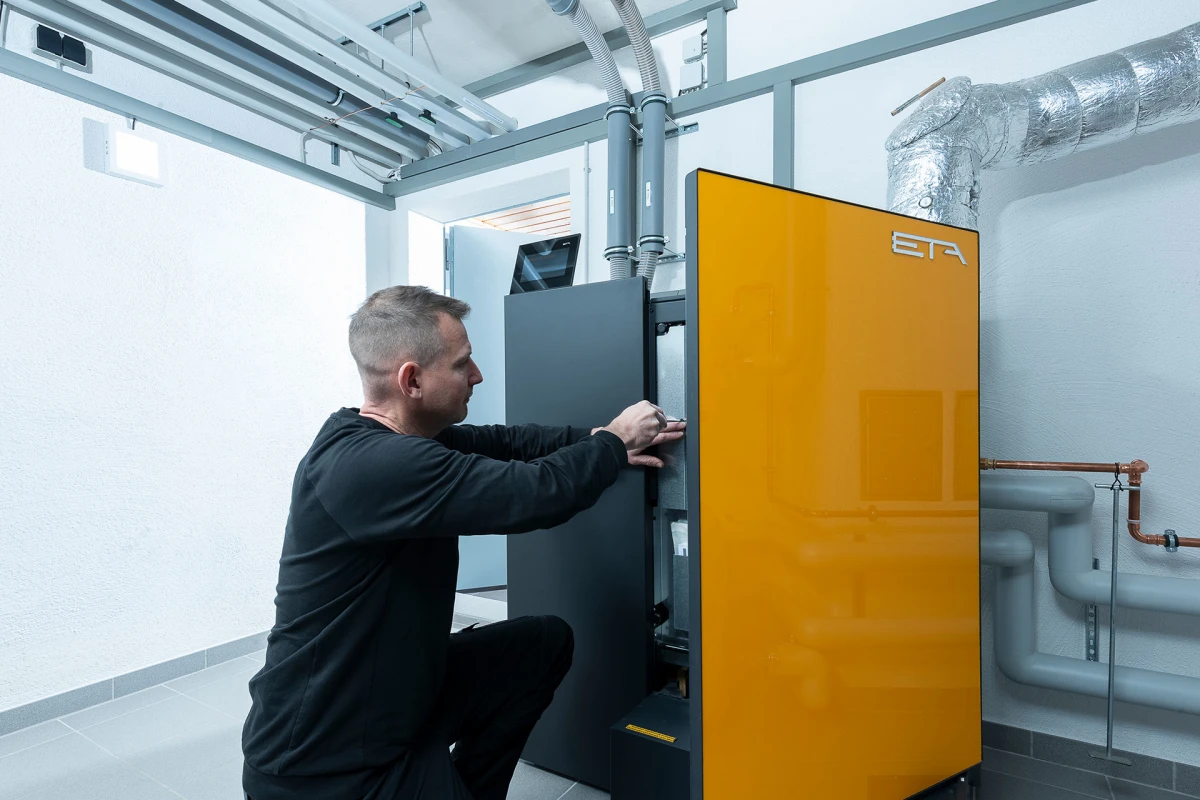 Ein Mann kniet neben einem großen orangefarbenen Heizkessel oder Industriegerät und justiert oder repariert dessen schwarze Seitenklappe in einem technischen Raum mit Rohren an der Decke.
