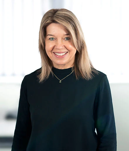 Frau mit schulterlangem blondem Haar, die ein schwarzes Shirt und eine zarte Halskette trägt und freundlich lächelt, vor einem verschwommenen hellen Hintergrund.