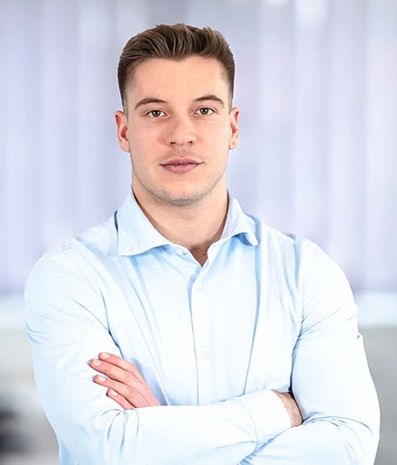 Junger Mann mit hellblauem Hemd steht mit verschränkten Armen vor verschwommenem Hintergrund und blickt direkt in die Kamera.