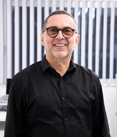Ein lächelnder Mann mit kurzen grauen Haaren und Brille trägt ein schwarzes Hemd vor einem Fenster mit vertikalen Jalousien.