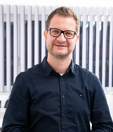 Ein lächelnder Mann mit kurzen braunen Haaren, schwarzer Brille und dunklem Hemd vor einem Hintergrund aus vertikalen weißen Lamellen.