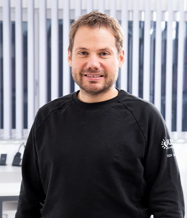 Porträt eines lächelnden Mannes mit kurzem braunem Haar und Bart, der ein schwarzes Sweatshirt trägt, vor einem Hintergrund mit weißen vertikalen Jalousien und einem weißen Schreibtisch.