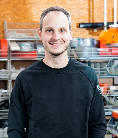 Ein lächelnder Mann mit kurzem braunem Haar trägt ein schwarzes Sweatshirt vor einem Regal mit Werkzeugen und Lagerbehältern in einer Werkstatt.