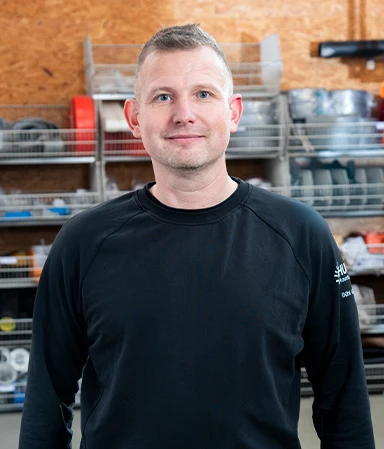 Mann mit kurzem hellbraunem Haar trägt schwarzen Pullover mit Logo auf dem linken Ärmel, steht in einer Werkstatt vor Regalen mit Werkzeug und Materialien.