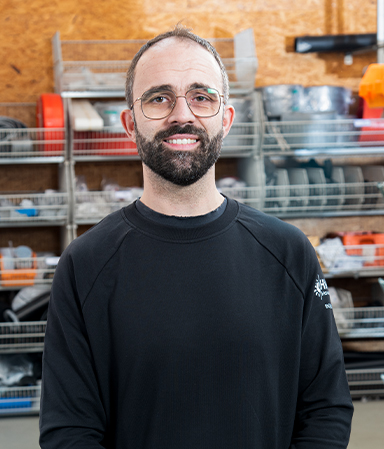 Mann mit Brille, Bart und kurzem Haar in schwarzem Pullover steht vor Regalen mit Werkzeugen und Materialien in einer Werkstatt.