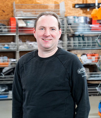 Porträt eines lächelnden Mannes mit hellen Haut und kurzen braunen Haaren, der einen schwarzen Pullover mit weißem Logo auf dem Ärmel trägt, vor einem Regal mit verschiedenen Werkstatt- und Industrieartikeln.