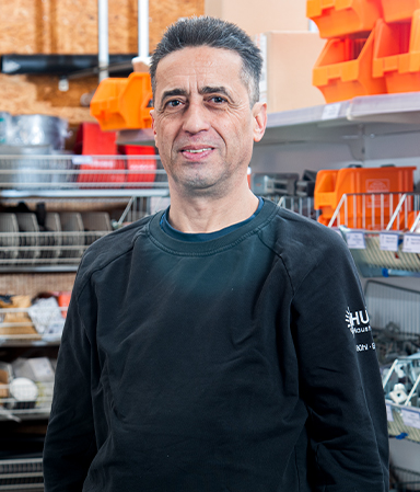 Mann mit dunklem Haar und schwarzem Pullover steht in einem Laden oder Lager mit Regalen voller Werkzeuge und orangefarbenen Aufbewahrungsboxen.
