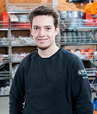 Junger Mann mit kurzem braunem Haar, Bart und schwarzen Pullover mit einem Firmenlogo auf dem Ärmel, steht lächelnd vor Regalen mit technischen Geräten und Ersatzteilen in einem Werkstatt- oder Lagerraum.