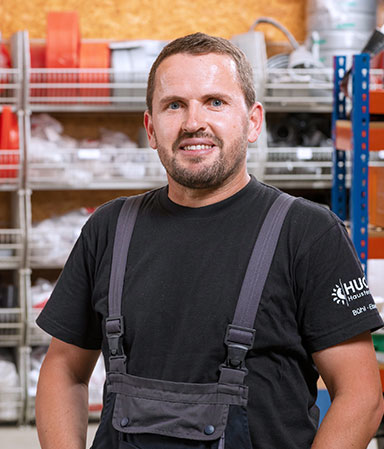 Ein Mann mit kurzem Haar und Bart trägt ein schwarzes T-Shirt mit Logo und graue Latzhose, stehend in einem Lager mit Regalen voller Werkzeuge und Materialien.