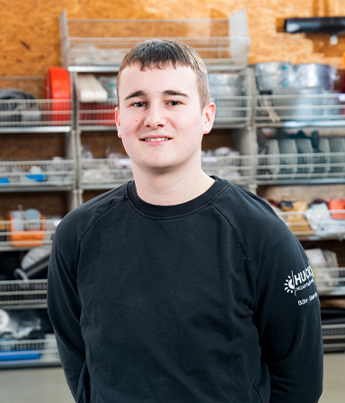 Junger Mann mit kurzem braunem Haar, der ein schwarzes Sweatshirt mit weißem Logo auf dem linken Ärmel trägt, steht vor Regalen mit Werkzeug und Ausrüstung in einer Werkstatt oder Garage.