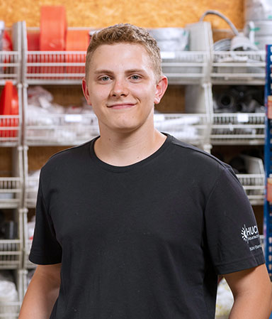 Ein junger Mann mit kurzen blonden Haaren trägt ein schwarzes T-Shirt und lächelt in einer Werkstatt mit Regalen voller Werkzeuge und Materialien im Hintergrund.