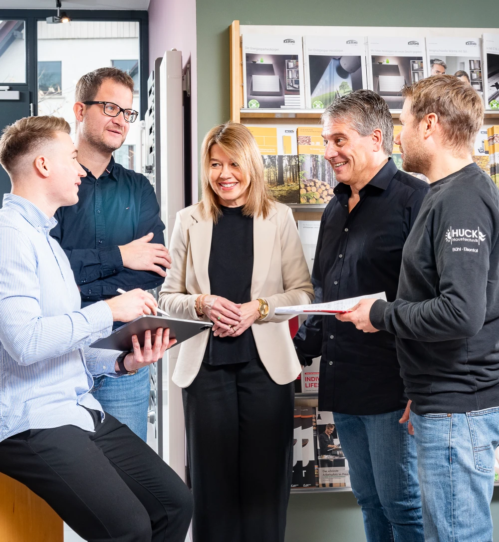 Fünf Personen in einem hellen Büro oder Geschäft, die lächelnd miteinander sprechen und Notizen machen, umgeben von Informationsbroschüren an der Wand.