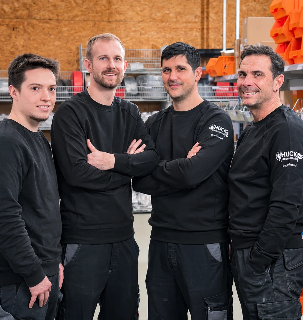 Vier Männer in schwarzer Arbeitskleidung mit dem Logo 'Huck Haustechnik' auf dem Ärmel, stehen lächelnd nebeneinander in einem Lagerraum mit Regalen und orangefarbenen Behältern im Hintergrund.