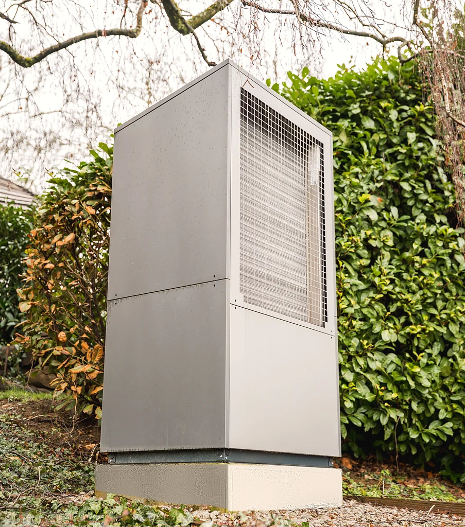 Eine freistehende, moderne Wärmepumpe mit grauer Metallverkleidung und Gitter auf der Vorderseite, installiert auf einem Sockel im Außenbereich mit grünem Buschwerk im Hintergrund.