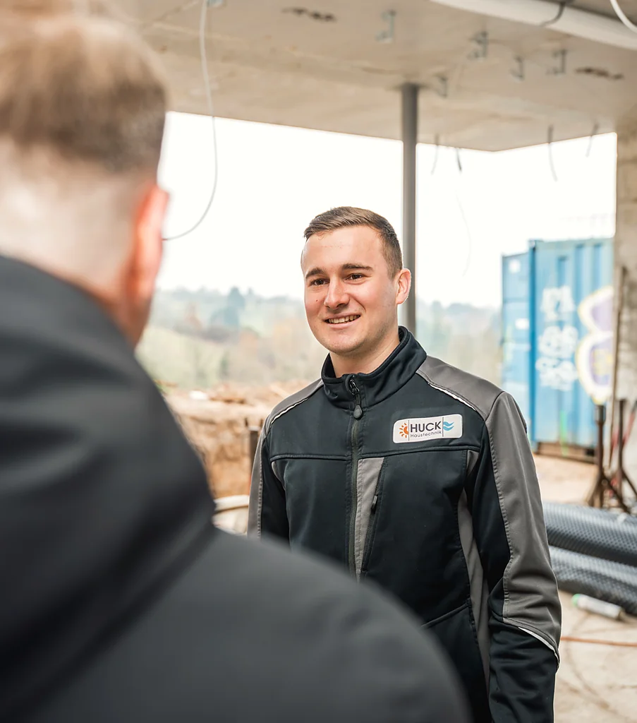 Zwei Männer auf einer Baustelle im Gespräch, einer von ihnen trägt eine Jacke mit dem Logo ‚HUCK Haustechnik‘ und lächelt.