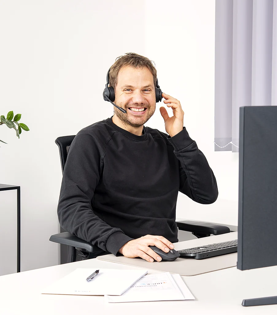 Ein lächelnder Mann mit Headset sitzt an einem Schreibtisch und benutzt eine Computermaus, vor ihm steht ein Monitor.