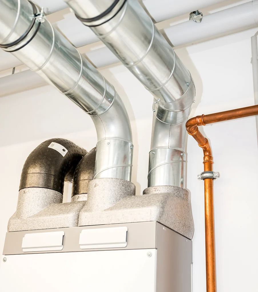 Moderne Lüftungsanlage mit silbernen Metallrohren und schwarzen Isolierstücken, daneben eine kupferfarbene Rohrleitung an einer weißen Wand.