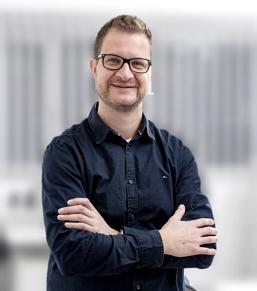 Mann mit Brille und schwarzem Hemd steht mit verschränkten Armen vor einem unscharfen, hellen Hintergrund und lächelt freundlich.